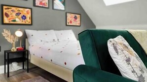 A bedroom with a bed, a green armchair, a small table with a lamp, and several framed artworks on the wall. - Bright Studio Loft 30min to Central London (Wembley)