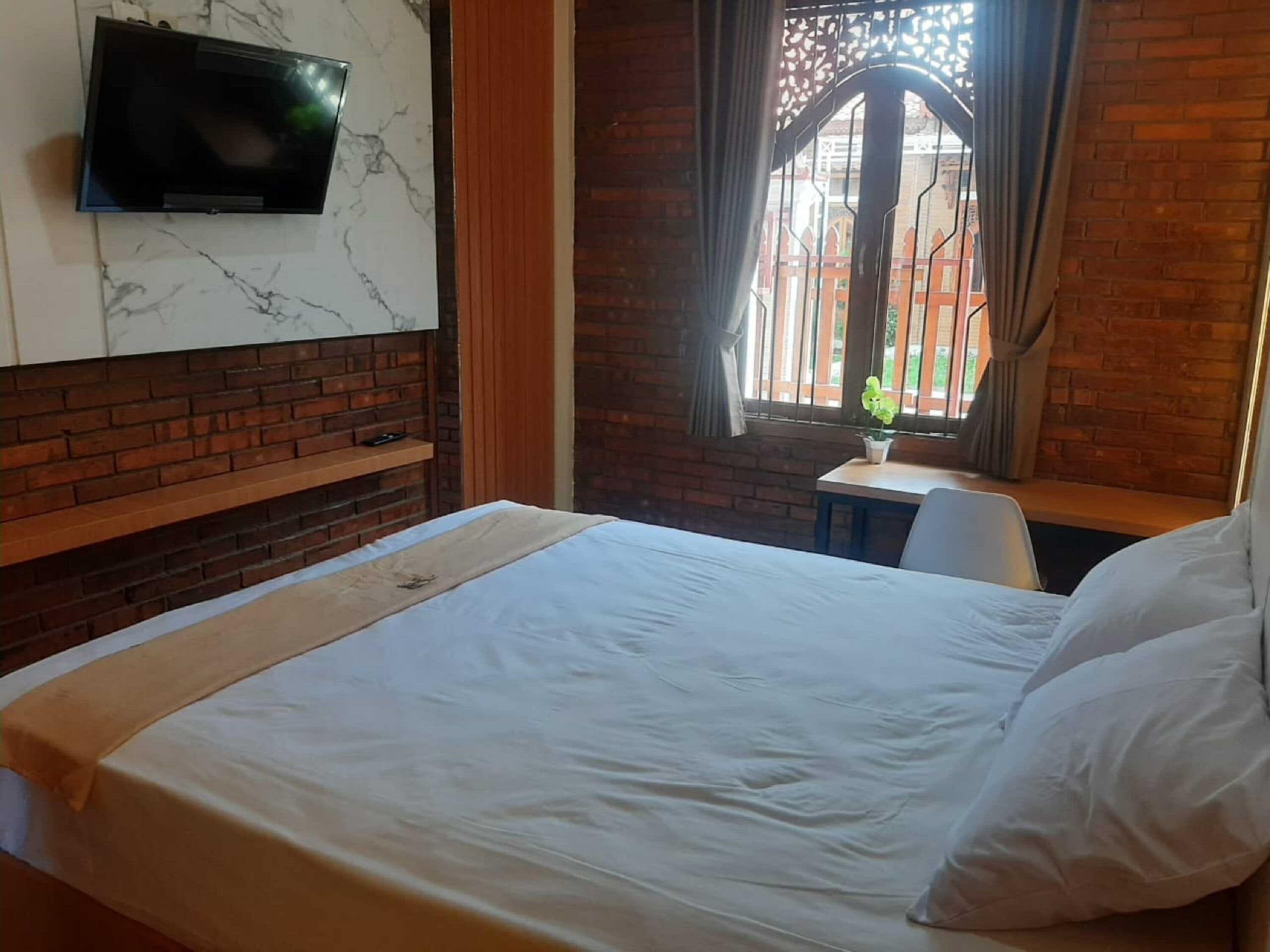 A hotel room with a bed, a TV mounted on a marble wall, a small table with a chair, and a window with a view of a building.