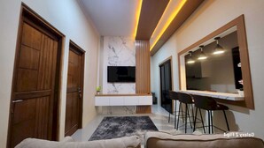 A modern living room with a sofa, a TV mounted on a marble wall, a dining area with a bar, and wooden doors.