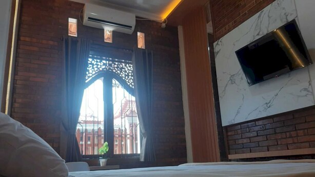 A hotel room with a bed, a TV mounted on the wall, a window with a decorative screen, and an air conditioning unit.