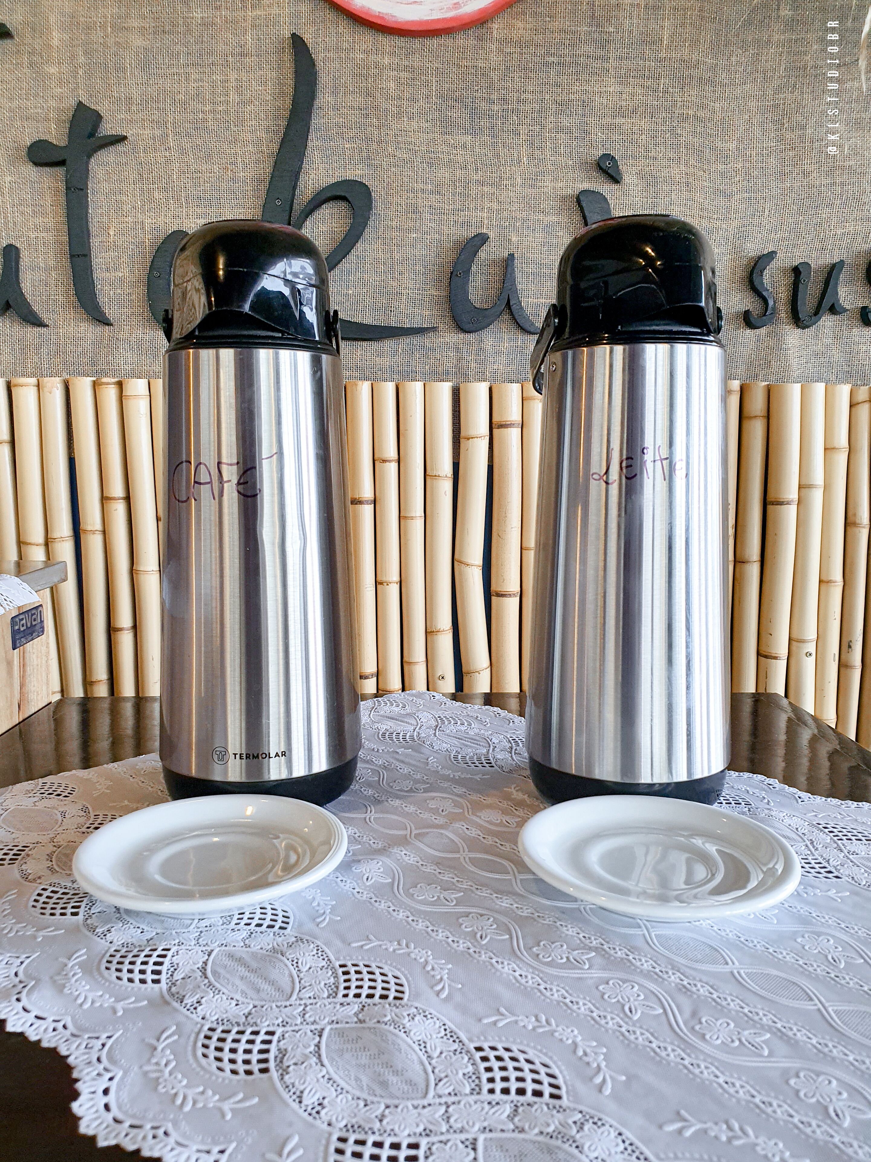 Two stainless steel thermos flasks on a table with a lace tablecloth, bamboo sticks, and a wall with a sign.
