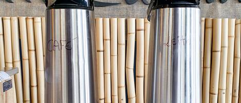 Two stainless steel thermos flasks on a table with a lace tablecloth, bamboo sticks, and a wall with a sign.