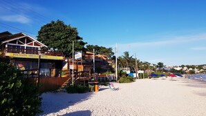 Um edifício à beira-mar com uma placa indicando 'Pousada Areia Cristal' e uma praia com guarda-sóis e pessoas.