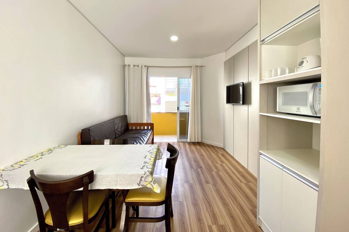 A small, well-lit room with a dining table and chairs, a sofa, a TV mounted on the wall, and a microwave on a shelf.