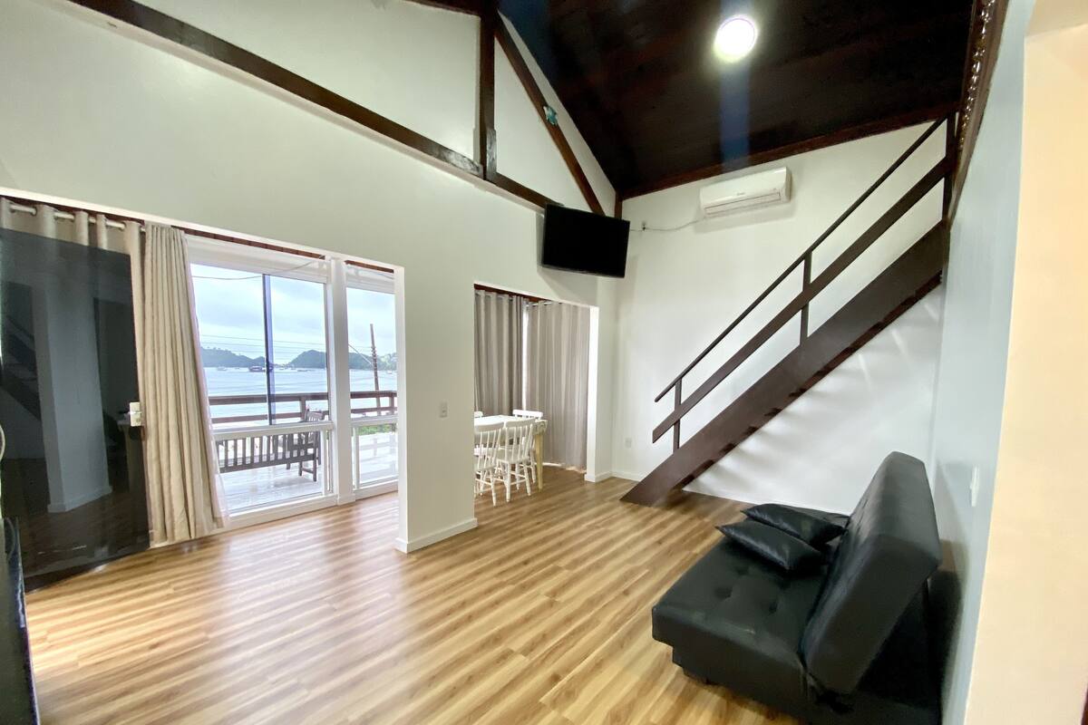 A spacious living area with a wooden floor, a black sofa, a staircase, and a balcony with a view of the sea.