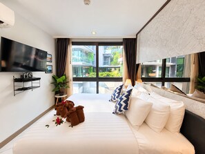 A hotel room with a large bed, a flat-screen TV, a small table with a plant, and a view of the outside.