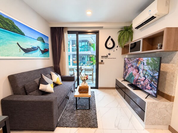 A modern living room with a grey sofa, a flat-screen TV, and a large window with a view of a building and greenery.