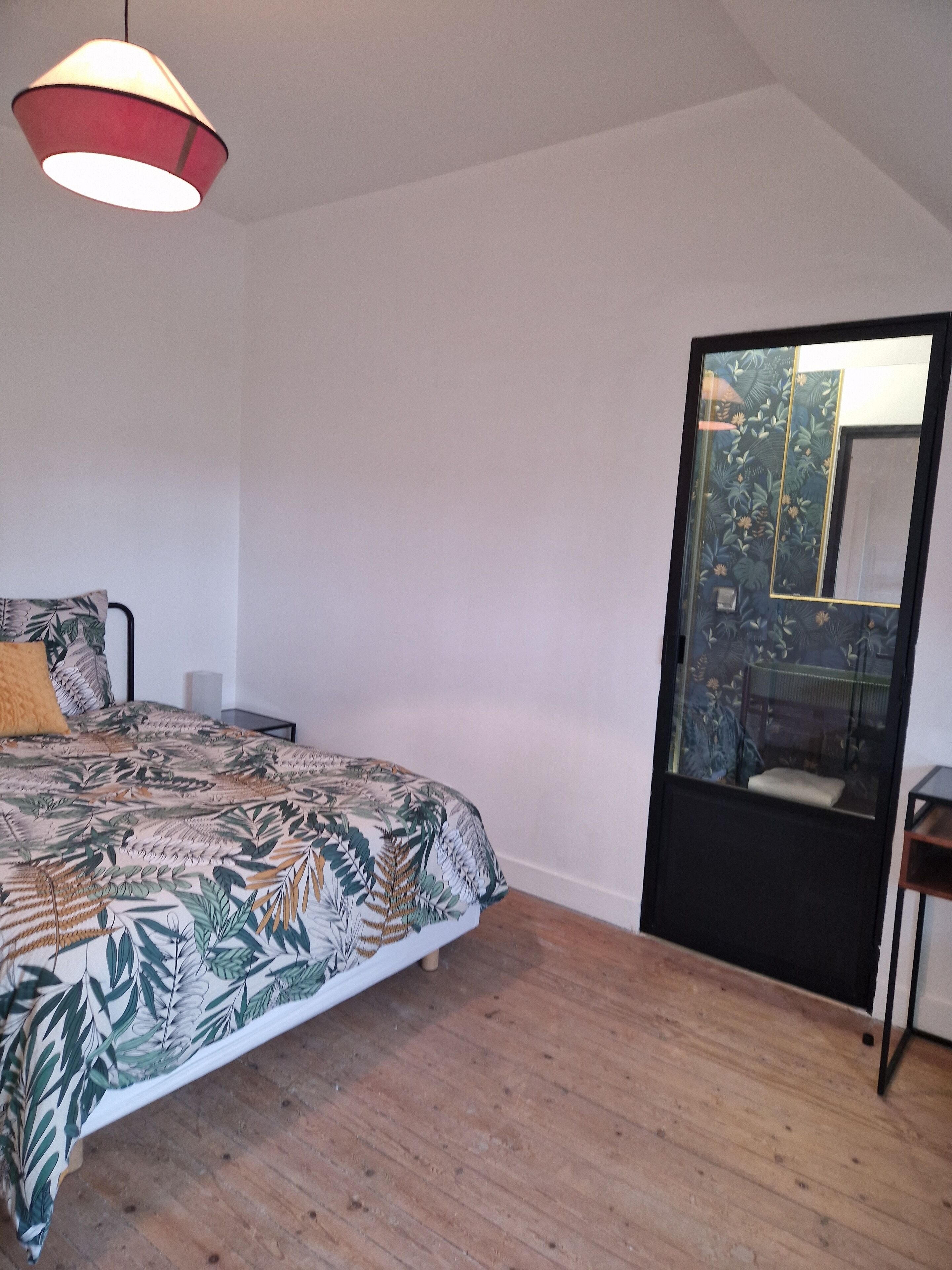 A bedroom with a bed, a nightstand, a wooden floor, a hanging light fixture, and a mirror with a floral pattern.