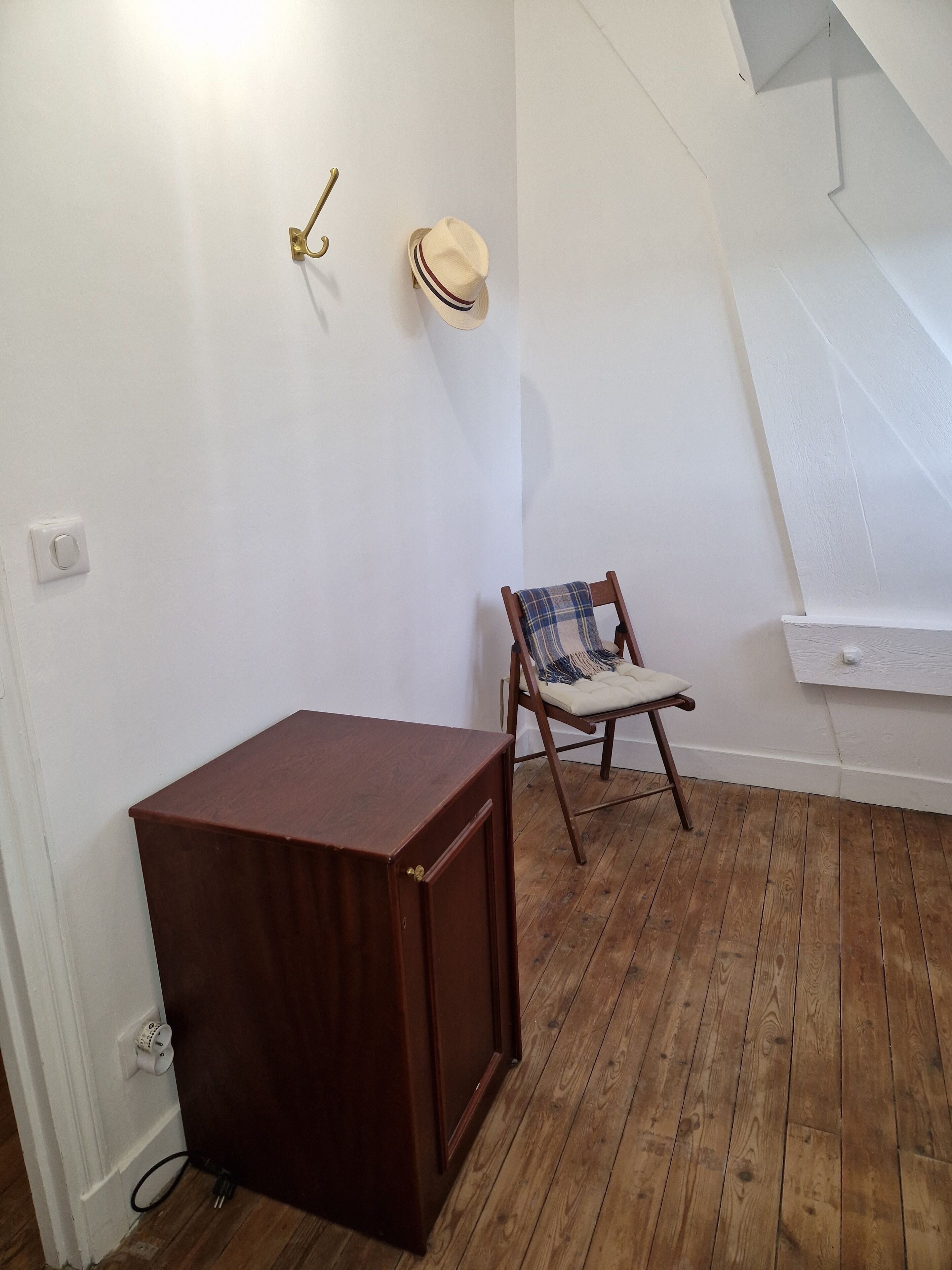 Una habitación con piso de madera, un mueble pequeño de madera, una silla con una manta de cuadros y un sombrero colgado en la pared.