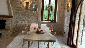 Une pièce rustique aux murs de pierre, avec une table à manger en bois, des lampes suspendues et une fenêtre donnant sur un paysage verdoyant.