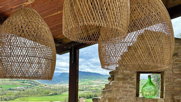 Un coin repas avec une table dressée pour quatre personnes, entouré de lampes suspendues en osier, offrant une vue sur la campagne et bordé d’un mur en pierre.