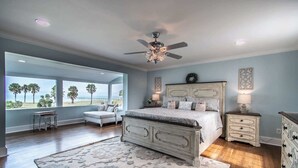 A bedroom with a large bed, a ceiling fan, a nightstand, a dresser, and a view of palm trees through large windows. - Coastal Oasis - Beautiful Ocean Views Awaits (Corpus Christi)