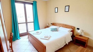 A bedroom with a bed, a nightstand, a wooden chest, a chair, and a sliding glass door with curtains. - LOCANDA DEL VIANDANTE (Magliano Alfieri)