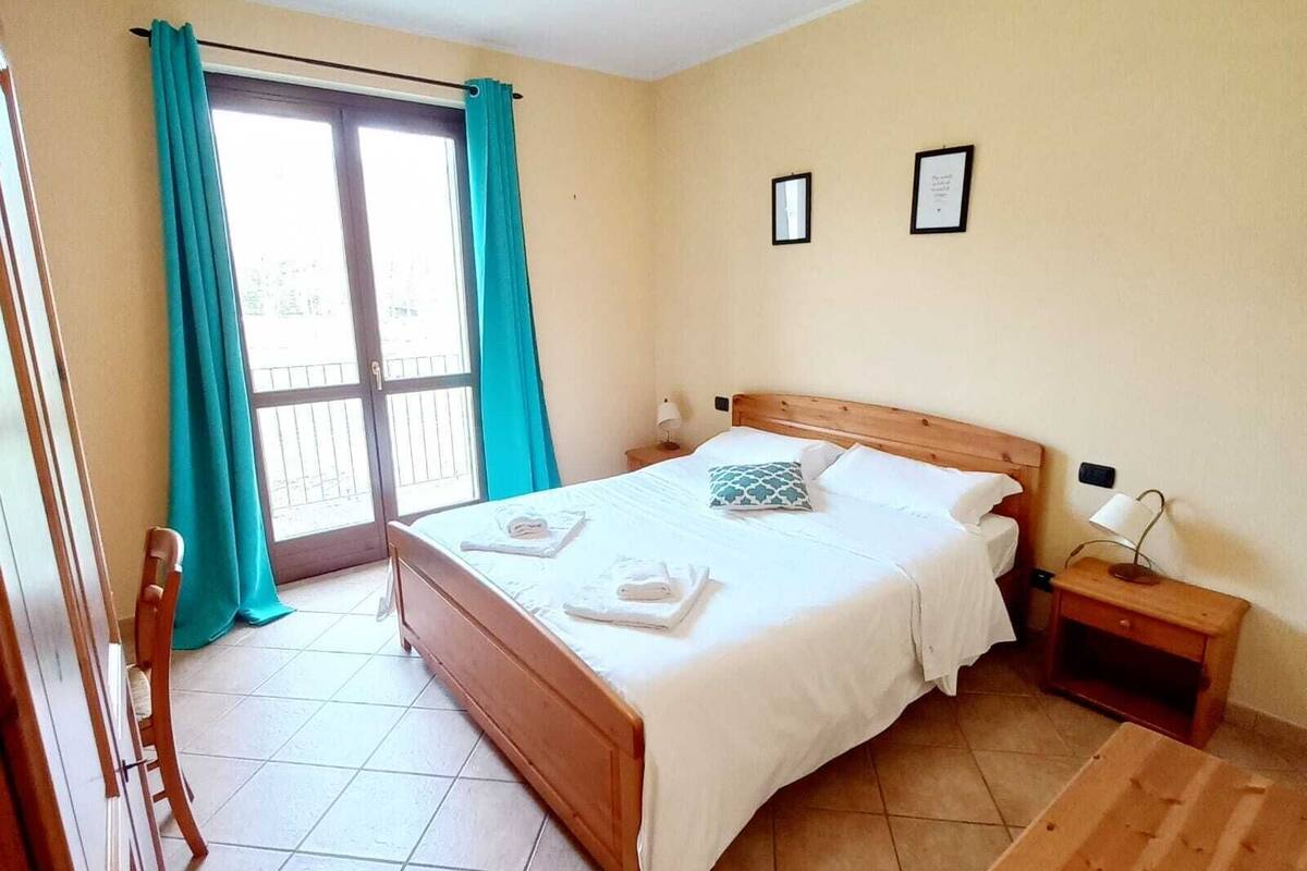Une chambre à coucher comprenant un lit, une table de chevet, une commode en bois, une chaise et une porte-fenêtre coulissante avec des rideaux.