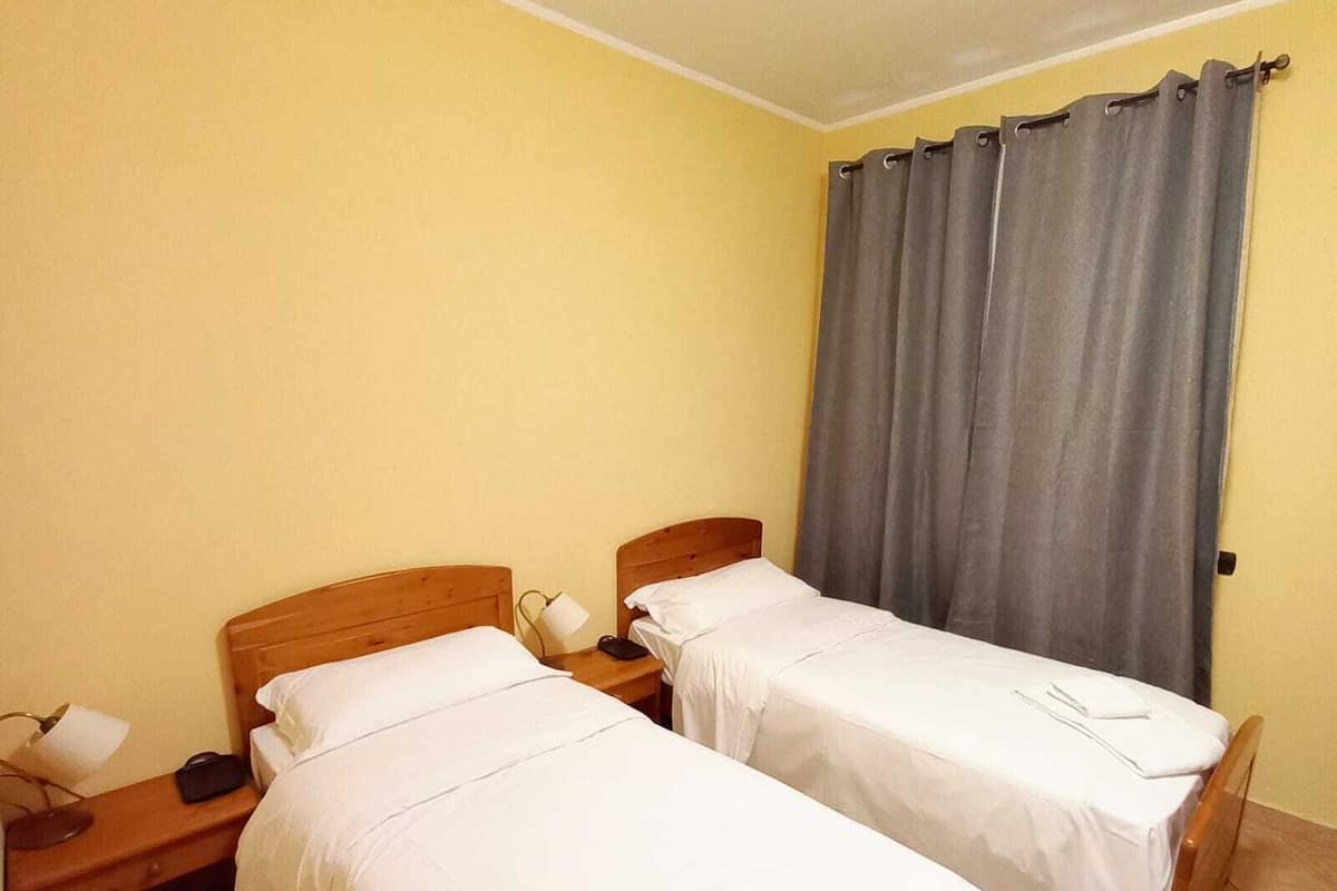 Une chambre d’hôtel avec deux lits simples, des têtes de lit en bois, du linge de lit blanc, une table de chevet avec une lampe et une fenêtre avec des rideaux gris.