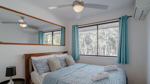 Une chambre à coucher comprenant un lit, une table de chevet, un ventilateur de plafond, une fenêtre avec des rideaux et un miroir.