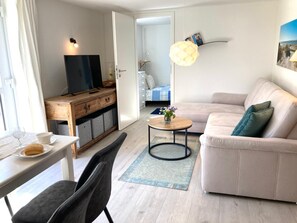 A modern living room with a sofa, a coffee table, a TV on a wooden cabinet, and a dining area with a table and chairs.