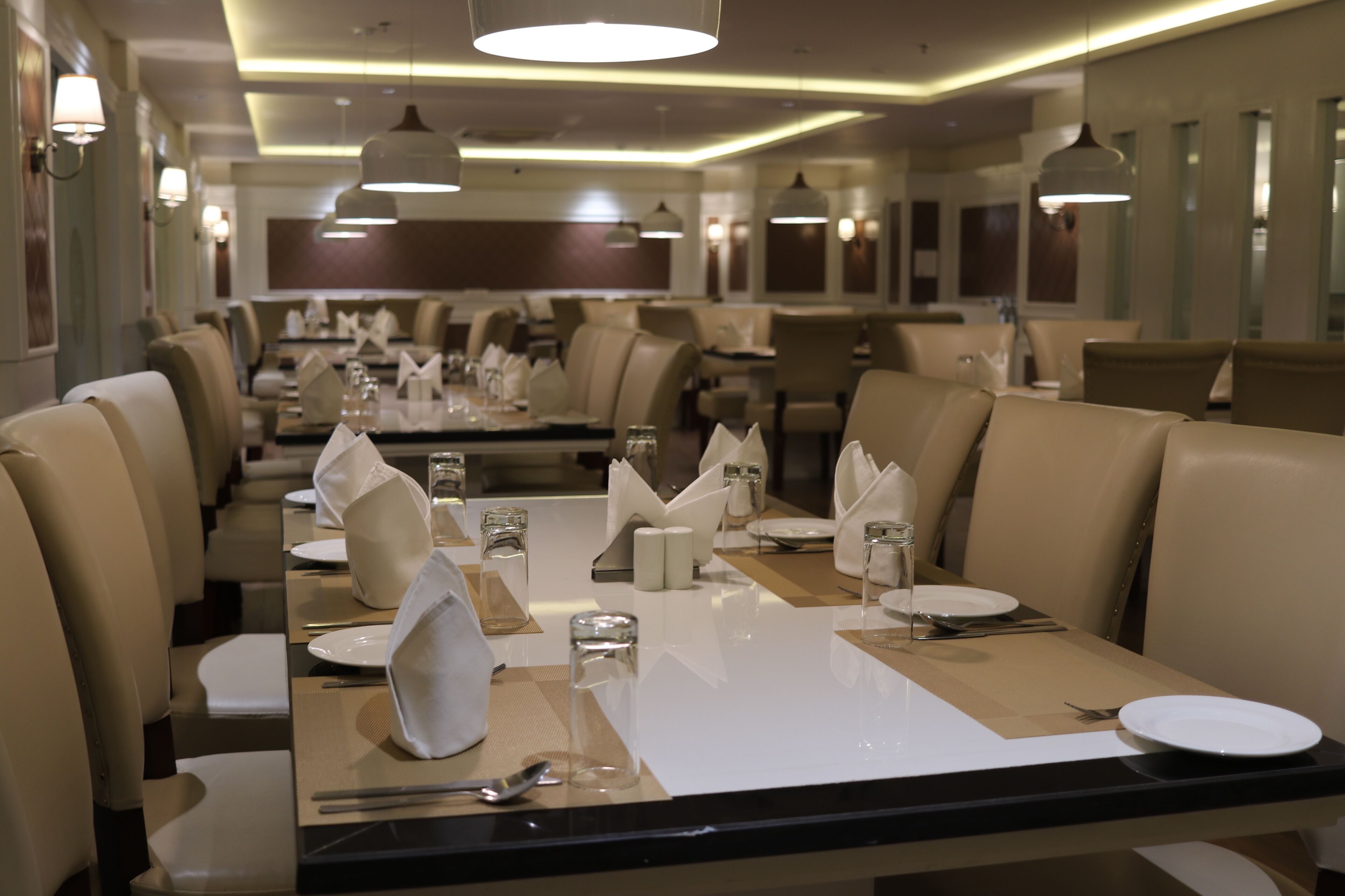Un restaurante con mesas dispuestas para comer, con un interior moderno que incluye iluminación colgante y una paleta de colores neutros.