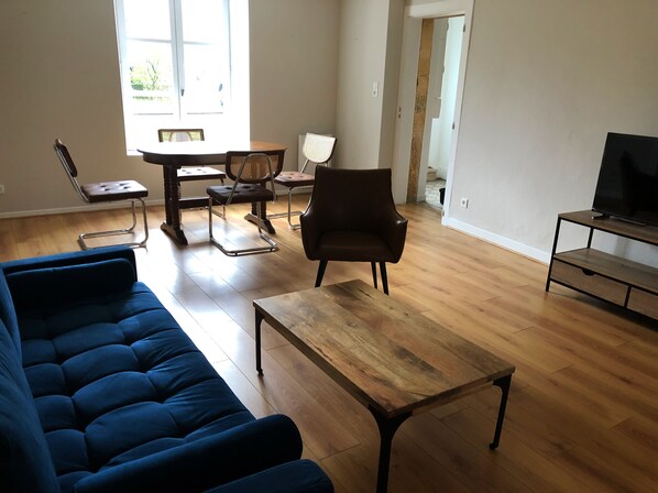 Een woonkamer met een blauwe bank, een houten salontafel, een tv-meubel en een eethoek met stoelen en een tafel.