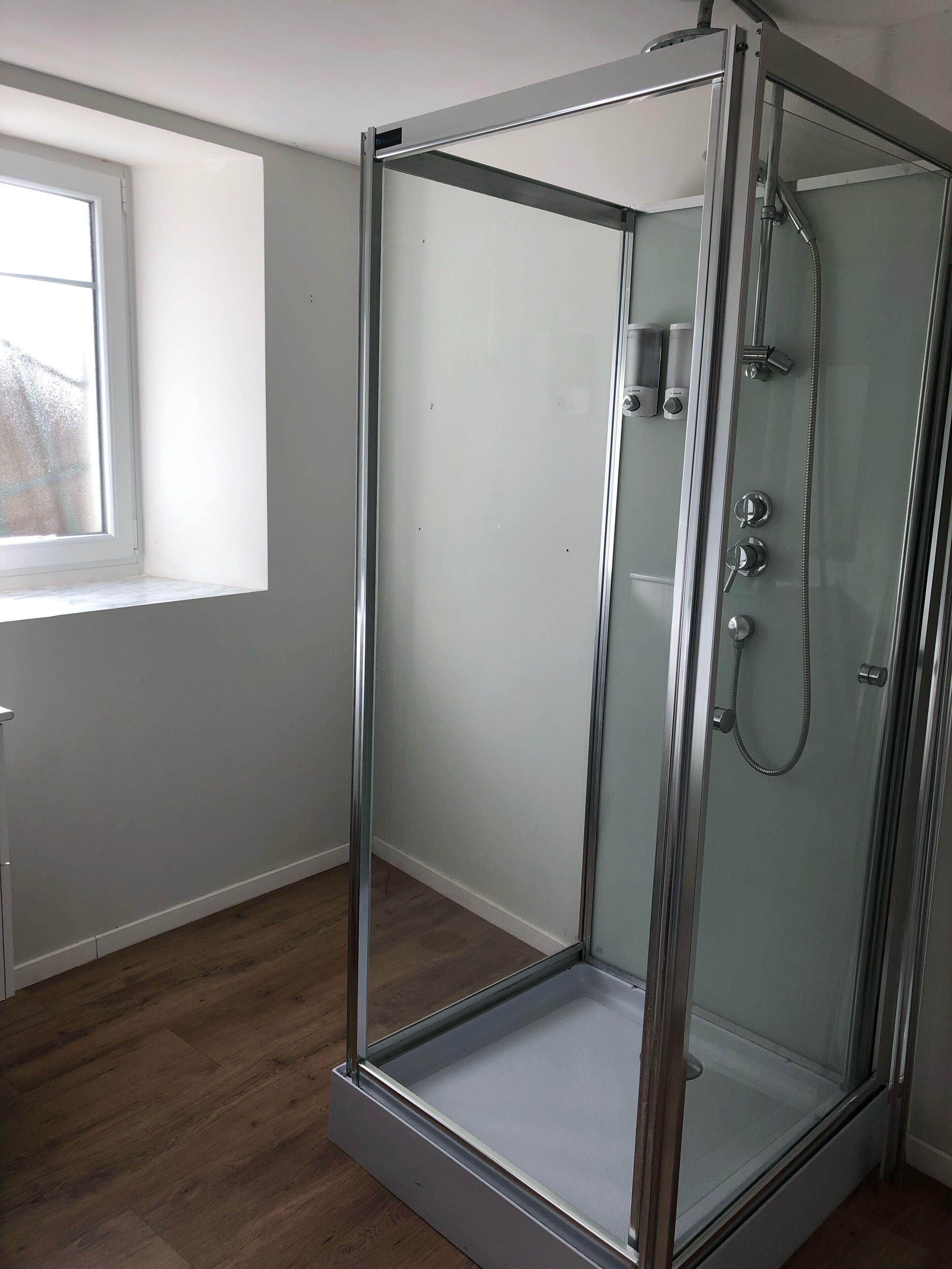 Une douche fermée par des parois de verre, avec une structure métallique, une fenêtre sur le côté gauche et un plancher en bois.
