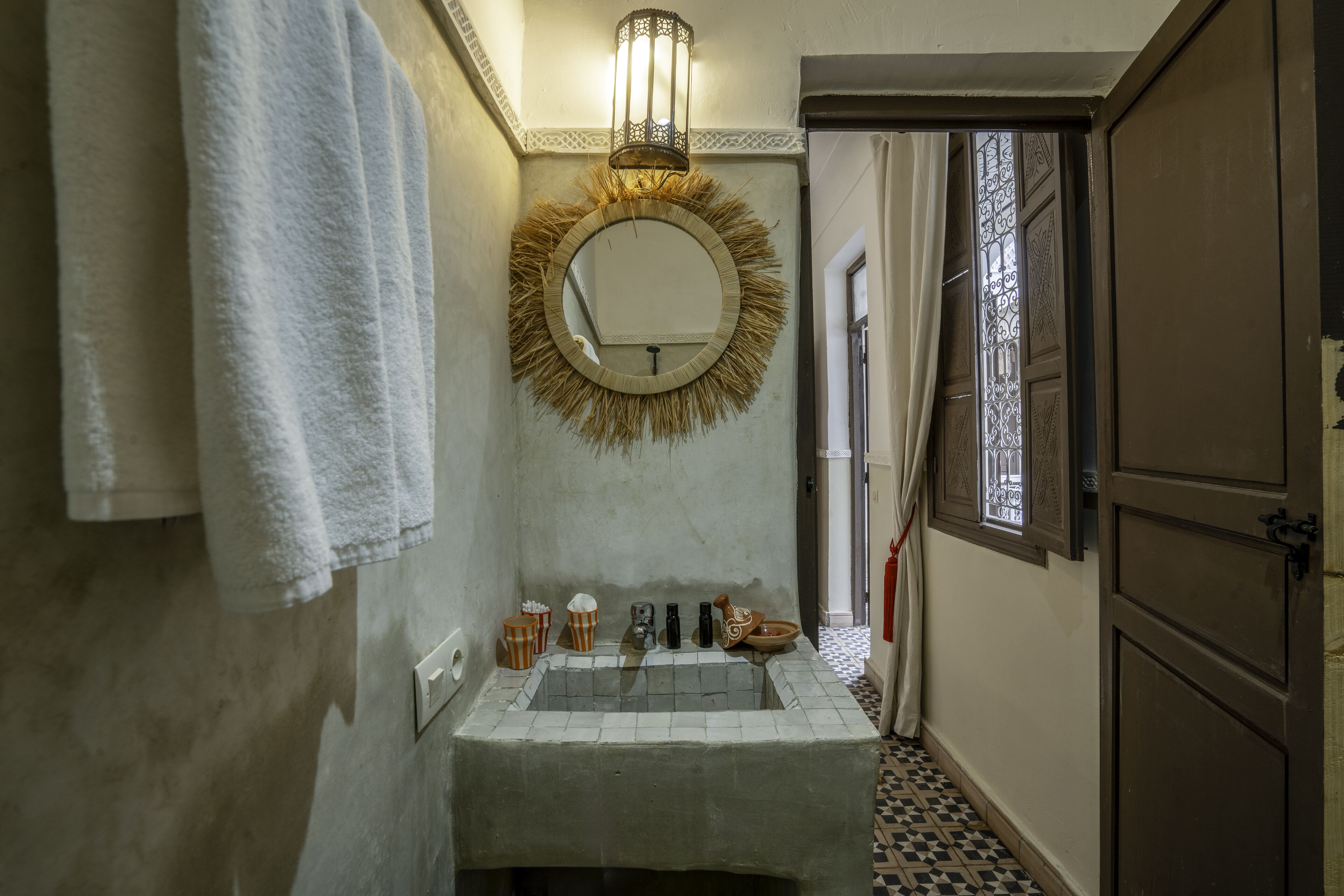 A bathroom with a round mirror, white tiles, a sink, and a window with a metal grill.