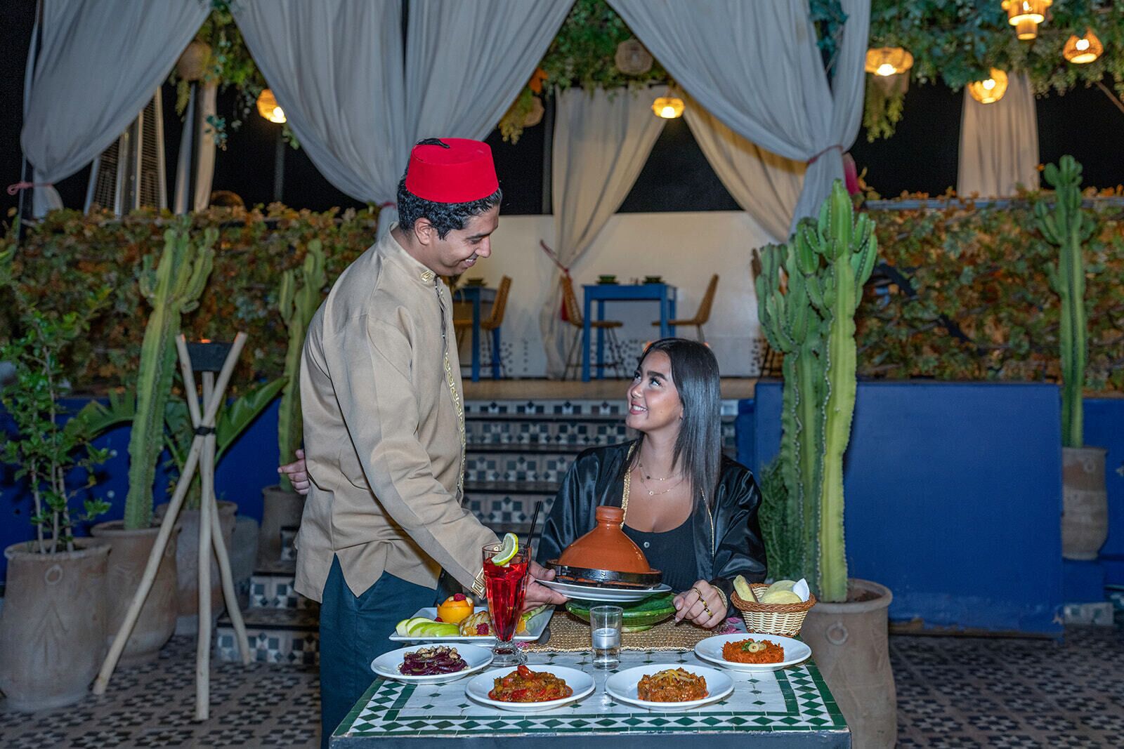Un homme, vêtu d’un costume traditionnel, sert des plats à une table en plein air, dont le sol est recouvert de carreaux et qui est agrémentée de plantes en pot.