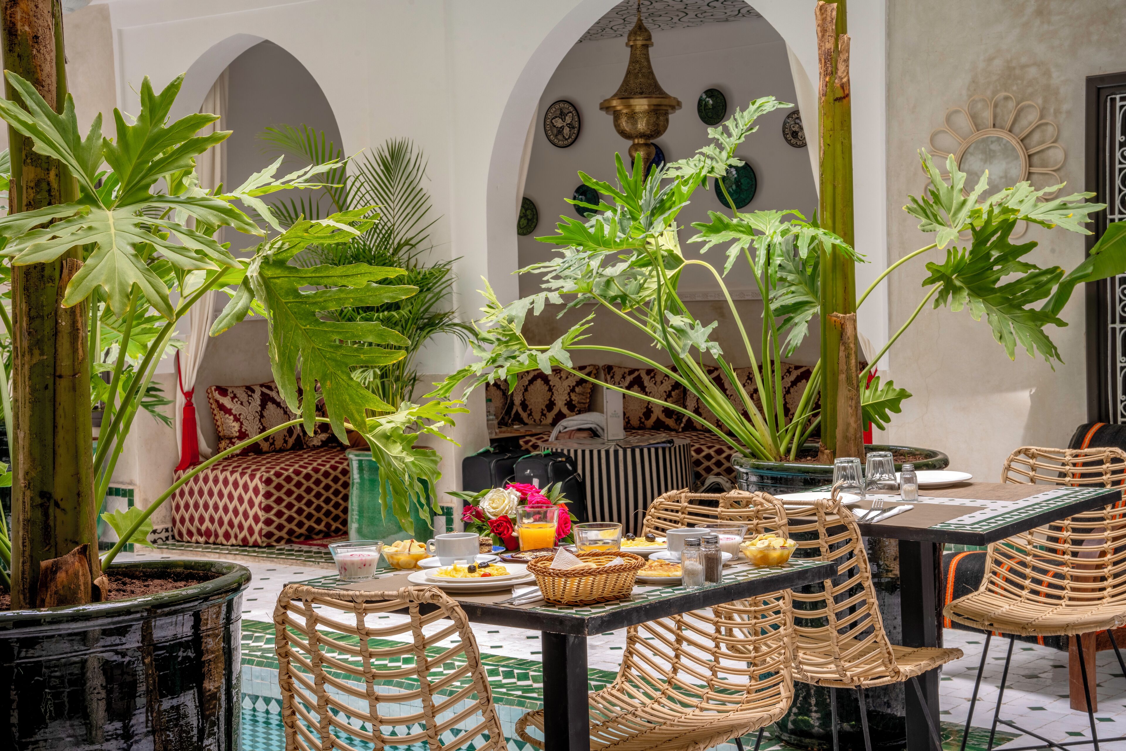 An outdoor dining area with wicker chairs and tables, surrounded by potted plants and a tiled floor.