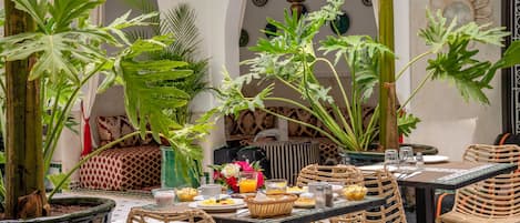 An outdoor dining area with wicker chairs and tables, surrounded by potted plants and a tiled floor.