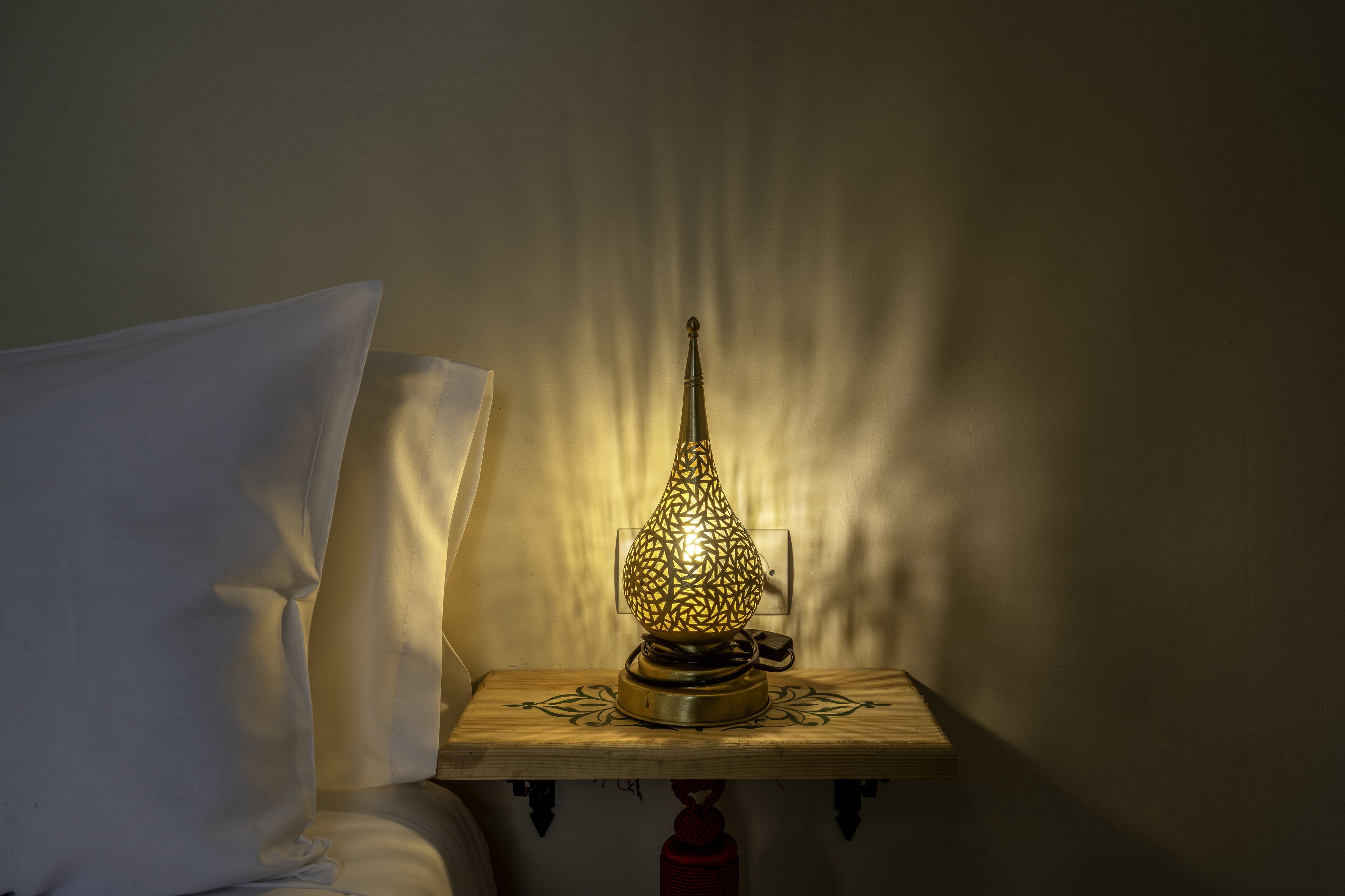 A bedside table with a decorative lamp, a pillow, and a wall-mounted light switch.