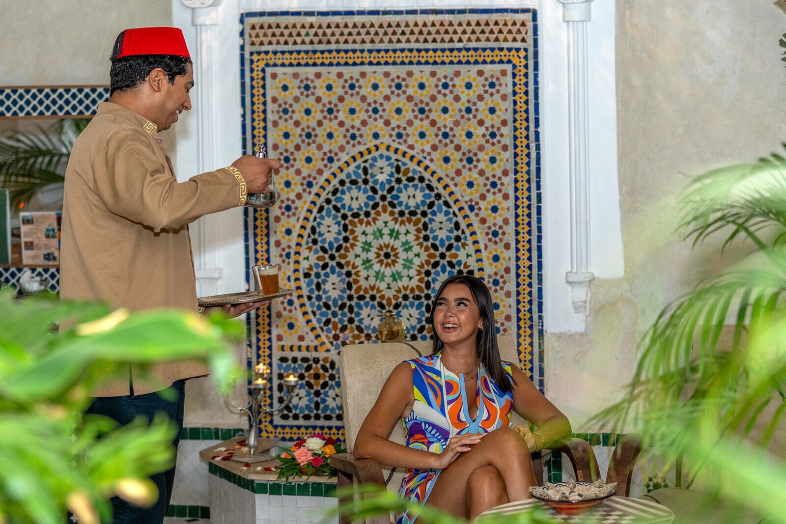 Una persona con vestimenta tradicional sirve una bebida a otra persona sentada en una habitación con un muro decorativo de azulejos.