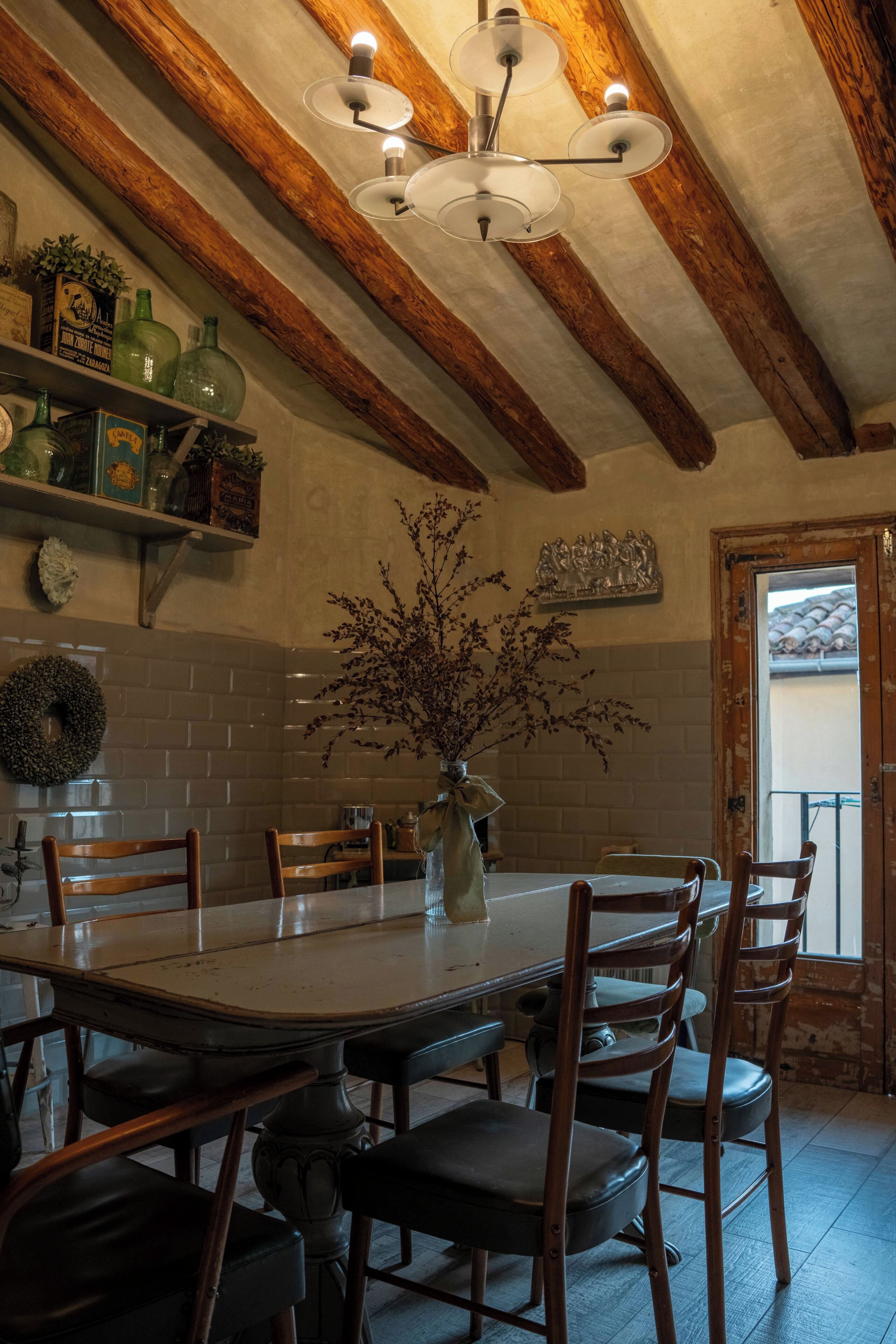 A dining area with a large wooden table, six chairs, a chandelier, and a vase with branches.
