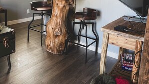 Un salon avec une table basse en bois au centre, deux tabourets de bar, un téléviseur posé sur un meuble en bois et un grand miroir au mur.
