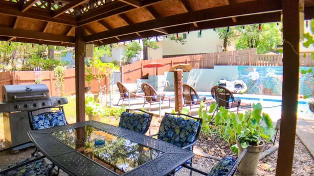 Une terrasse couverte avec une table et des chaises à plateau en verre, un barbecue et une piscine en arrière-plan.