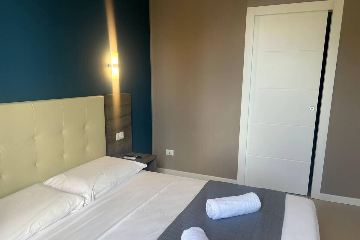 A hotel room with a large bed, a nightstand, a wall-mounted lamp, and a door.