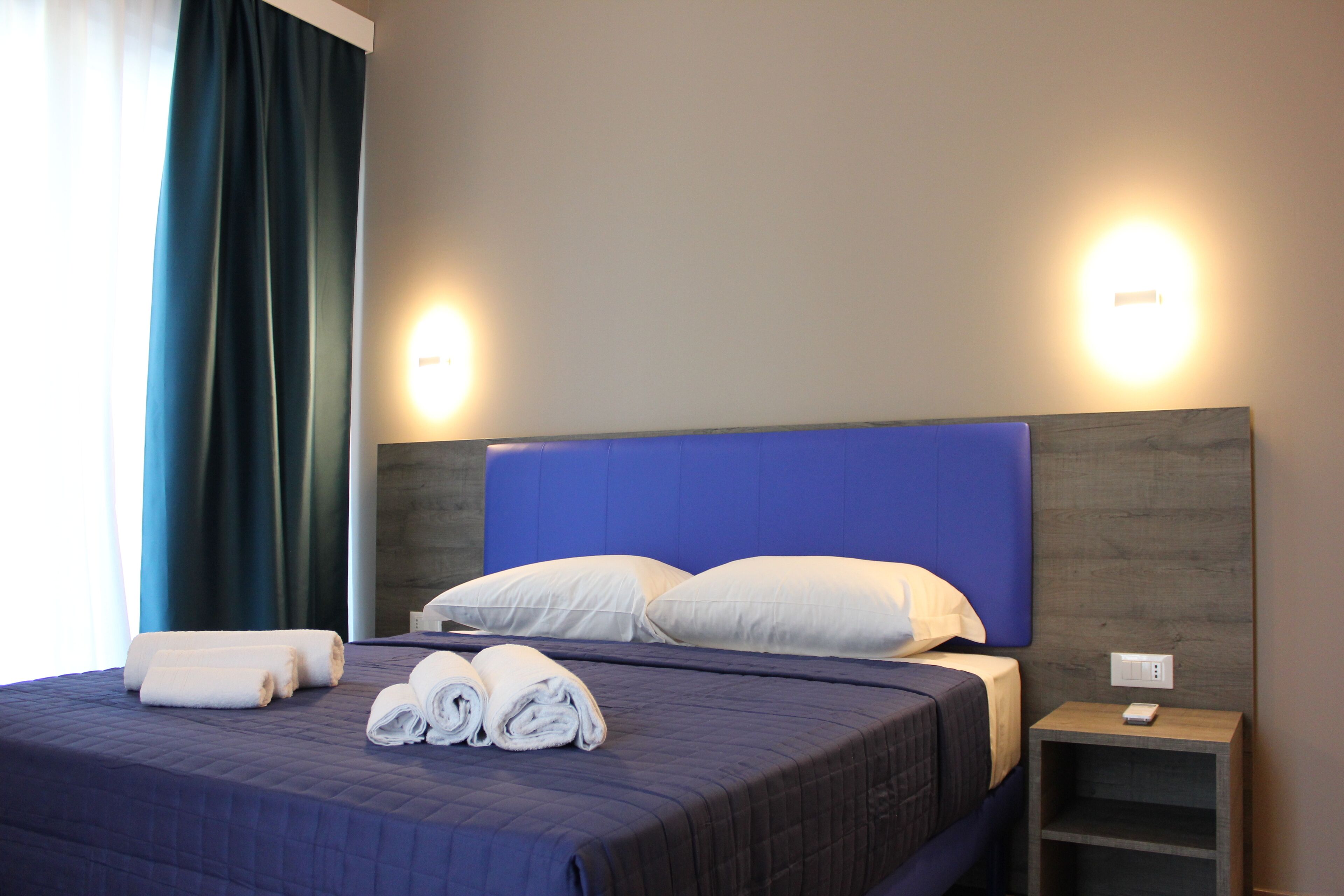 A neatly made bed with rolled towels, a blue headboard, and bedside lamps.
