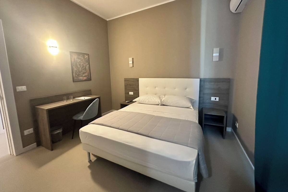 A bedroom with a large bed, a desk, and a chair. There is a wall-mounted light and a painting on the wall.