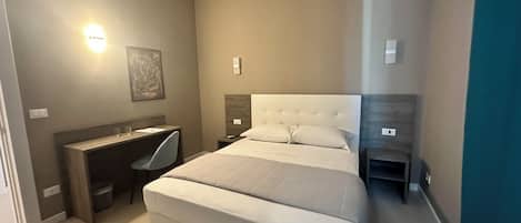 A bedroom with a large bed, a desk, and a chair. There is a wall-mounted light and a painting on the wall.