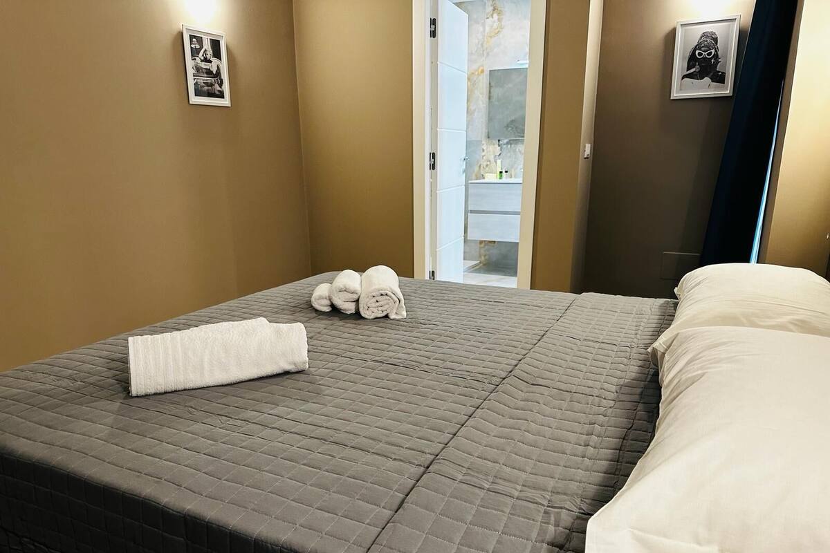 A neatly made bed with a quilted gray bedspread, two rolled white towels, and a white pillow.