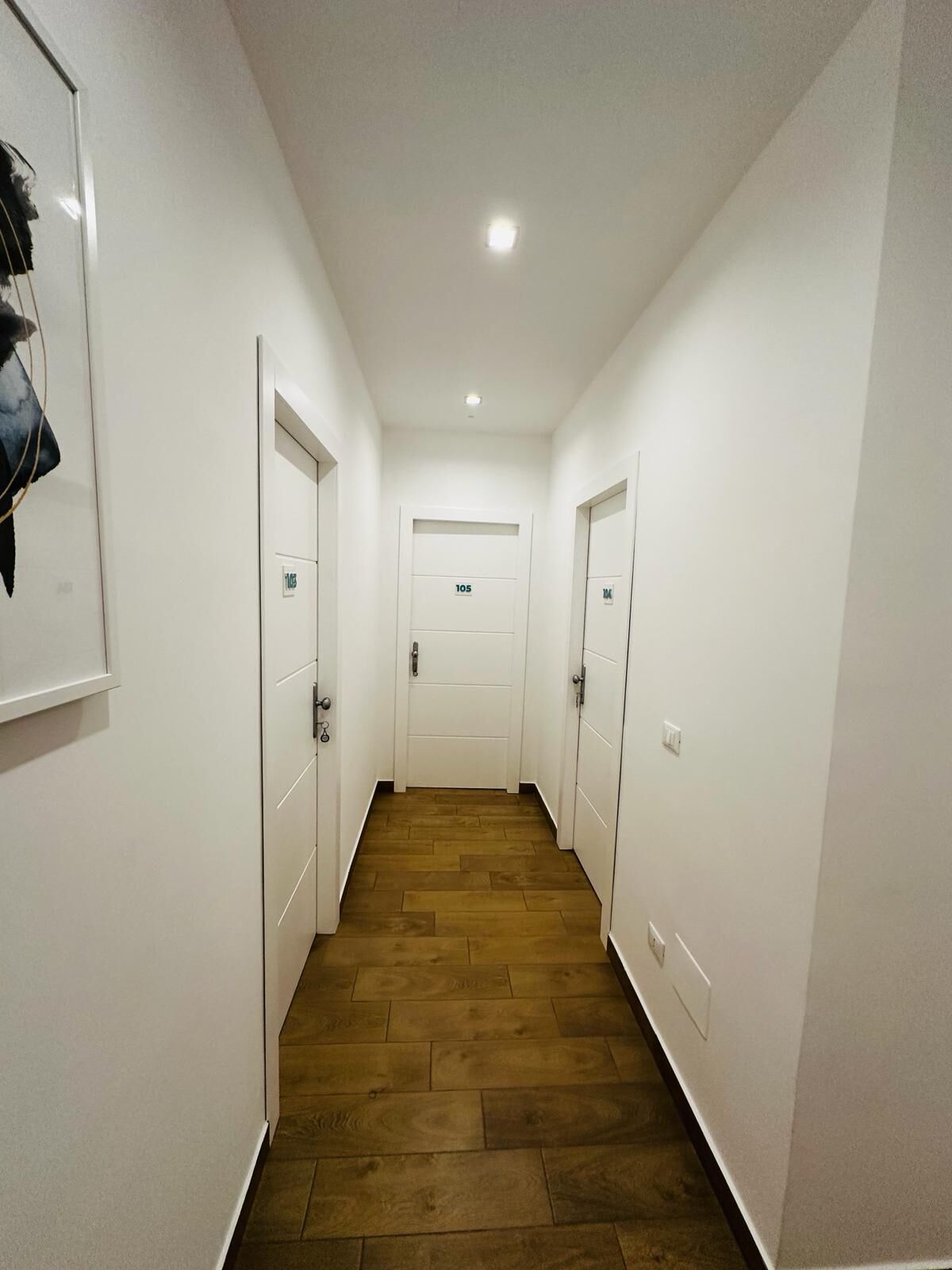 A narrow hallway with wooden flooring and white walls. There are three doors on the right side, each with a number above the door. The hallway has a ceiling light and a door at the end with a number above it.