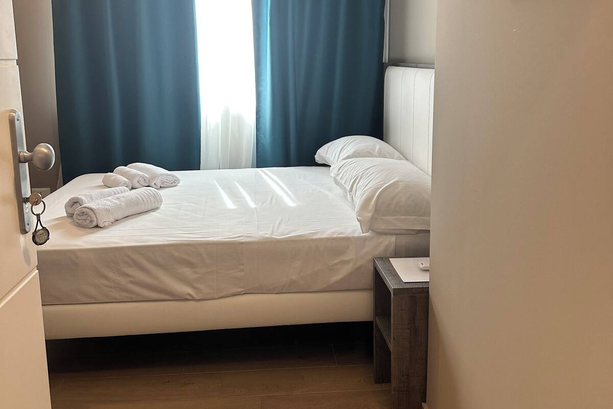 A hotel room with a bed, a nightstand, a framed picture on the wall, and a window with curtains.