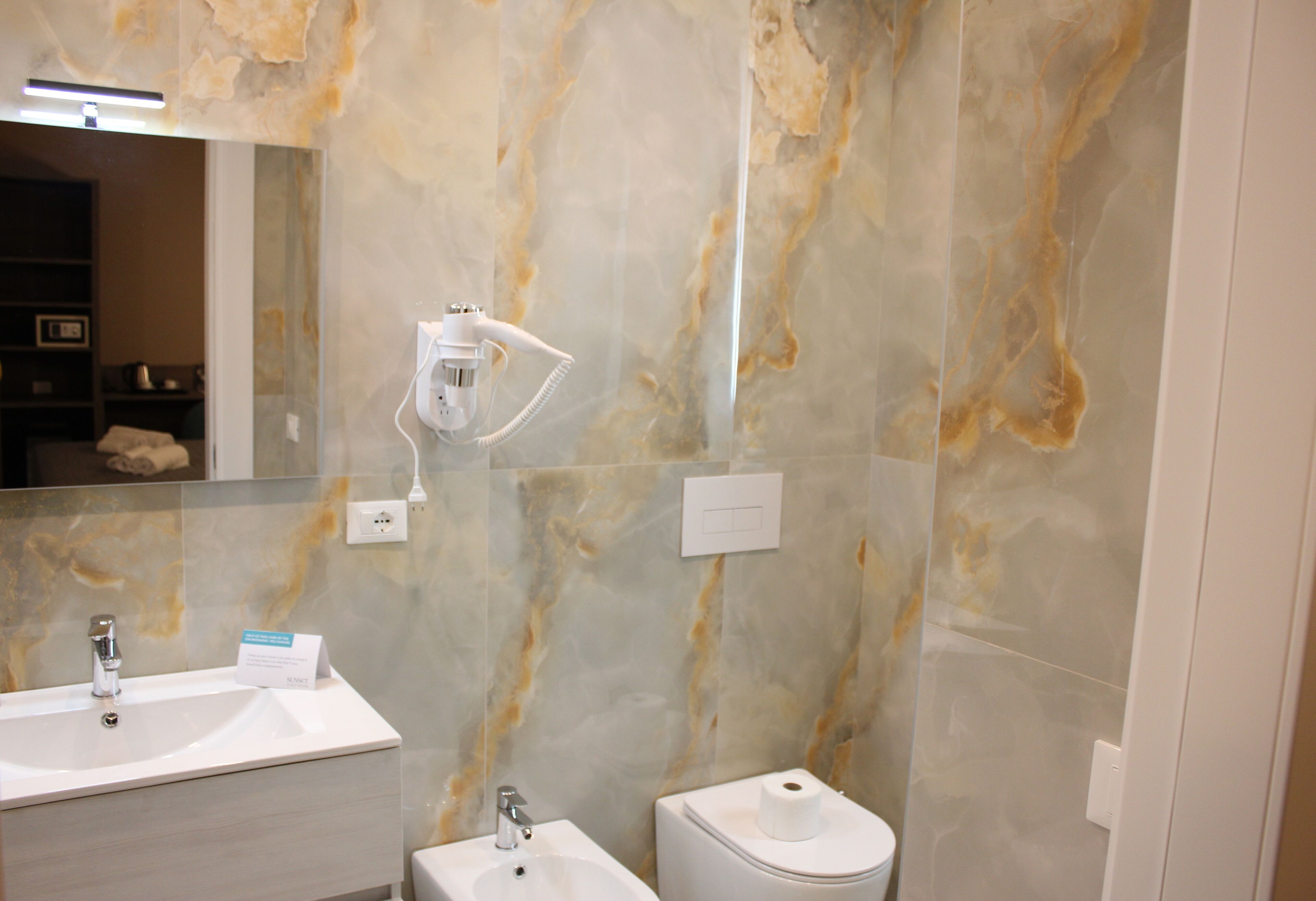 A bathroom with a marble wall, a sink, a mirror, a handheld showerhead, and a toilet.