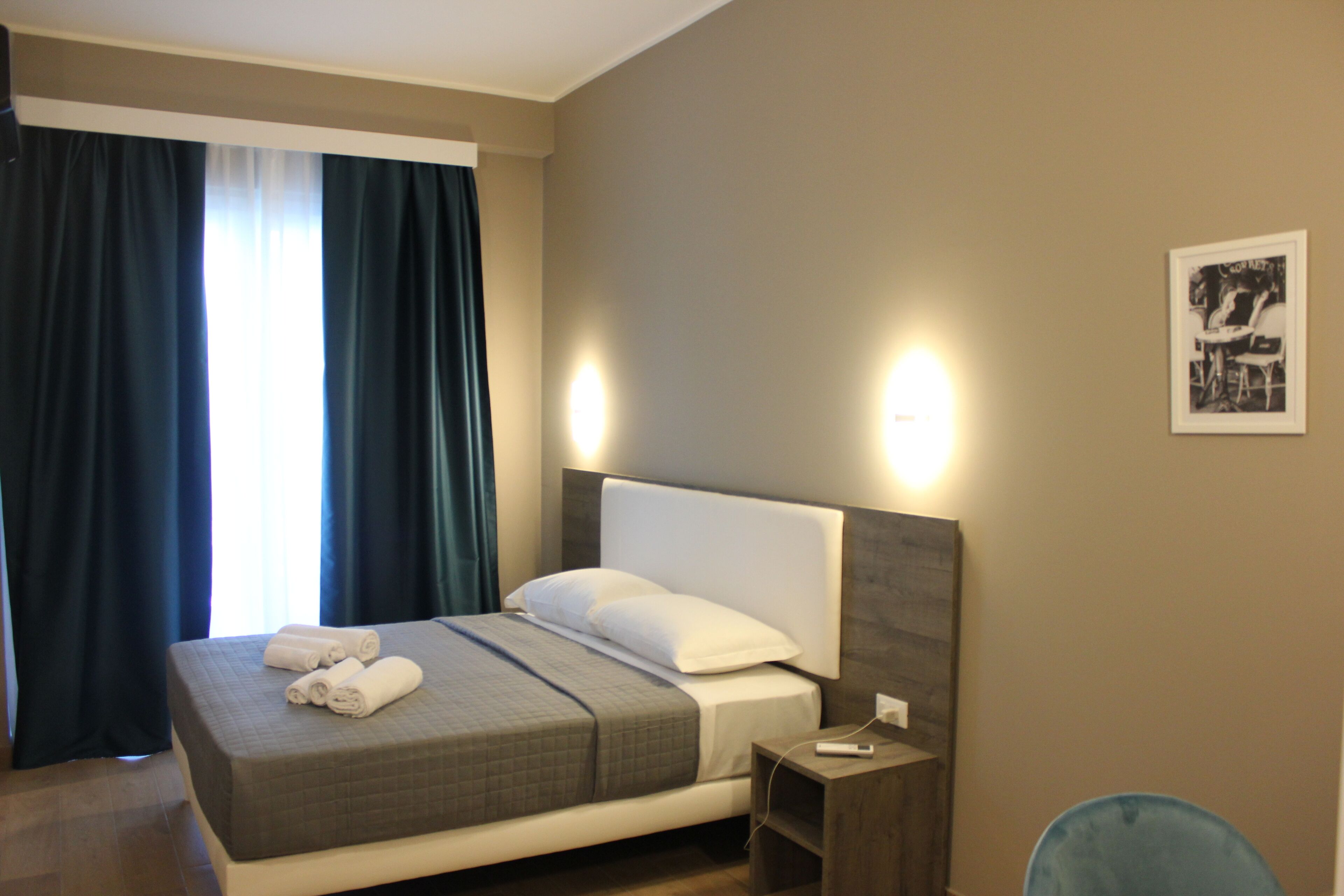 A neatly made bed with a grey quilt, white pillows, and rolled towels on the side. There is a bedside table with a lamp and a small nightstand.