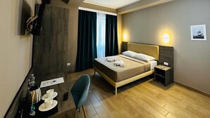 Et hotelværelse med seng, natbord, skrivebord med stol, fjernsyn og et vindue med gardiner.