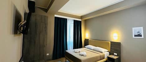 Habitación de hotel con cama, mesita de noche, escritorio con silla, televisor y ventana con cortinas.