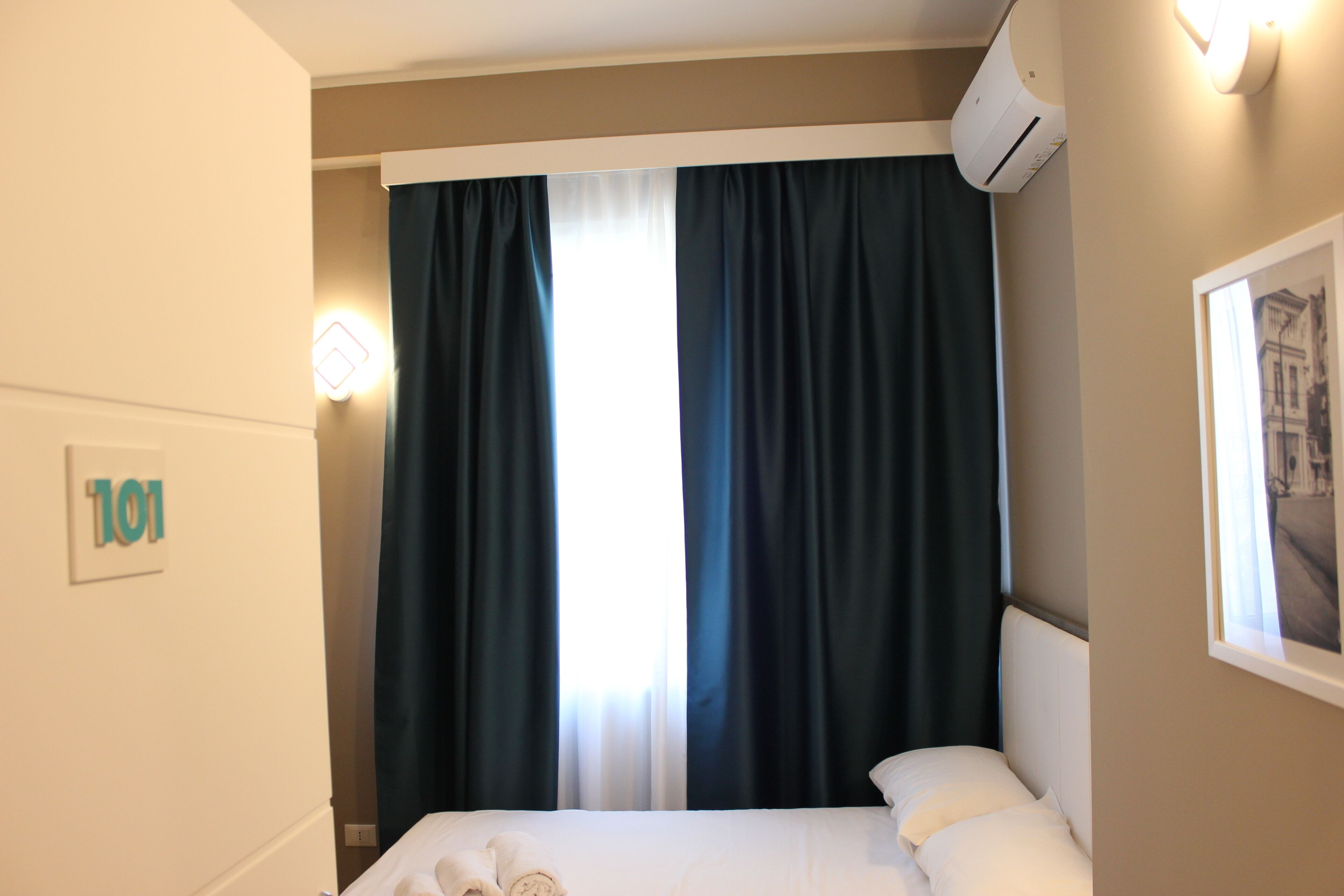 Habitación de hotel con cama, ventana con cortinas oscuras, unidad de aire acondicionado y un cuadro en la pared.