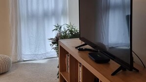 A living room with a flat-screen TV on a wooden stand, a bookshelf filled with books, a potted plant, and a large window with sheer curtains. - Stunning 2-bed Apartment Near Colindale Station (London)