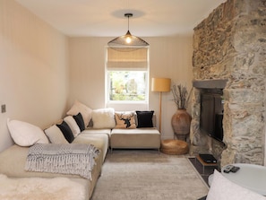 A cozy living room with a stone fireplace, a sofa set, a hanging light fixture, and a view through a window. - Hendre (Menai Bridge)