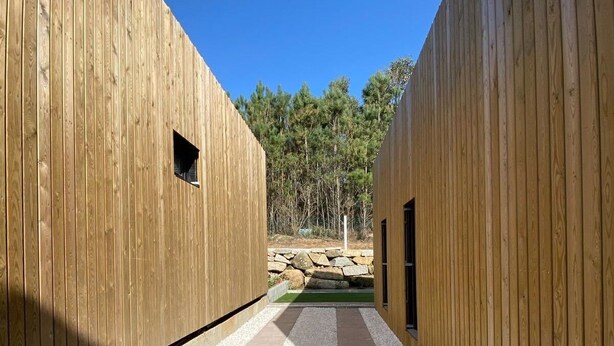 A corridor with wooden walls and a gravel path leading to a stone wall and trees. - Top Balea (O Grove)
