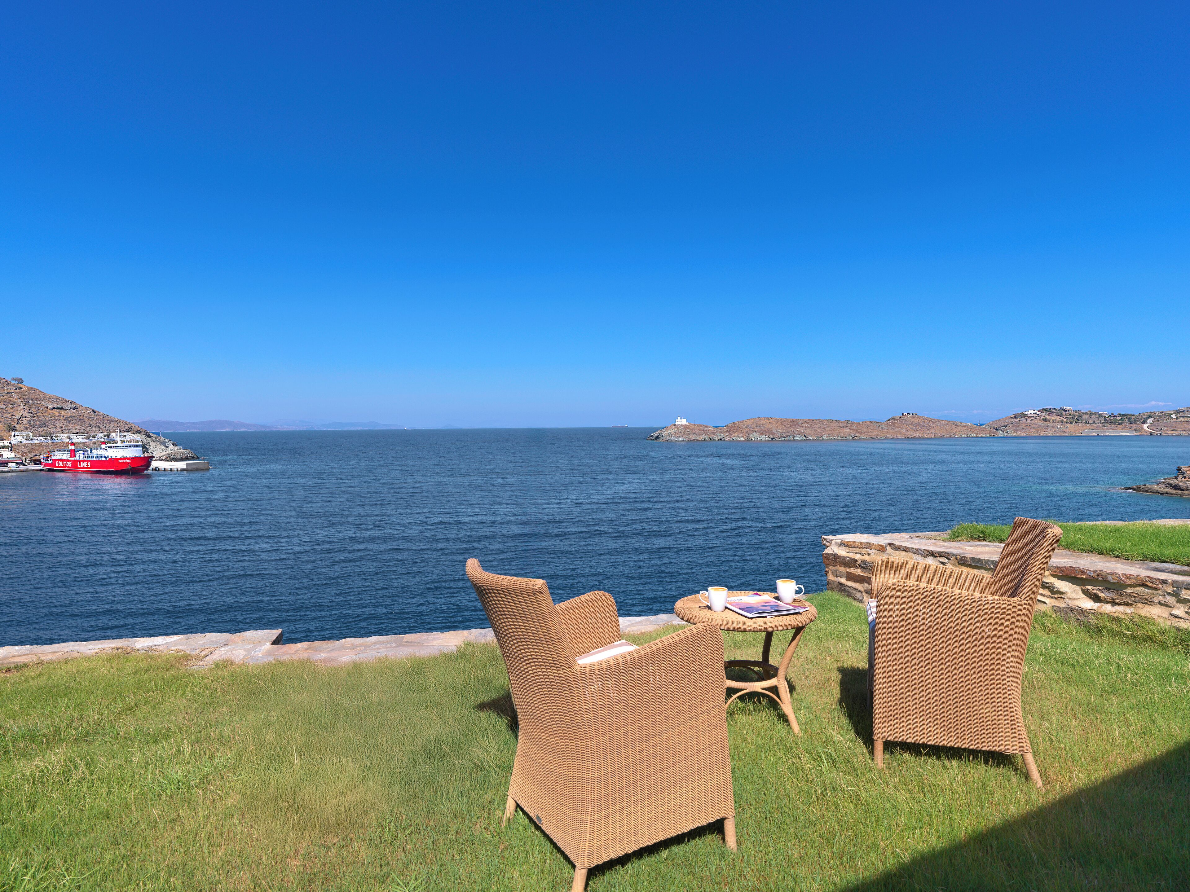 A seaside view with wicker chairs and a table, a red boat in the distance, and a lighthouse on a hill.