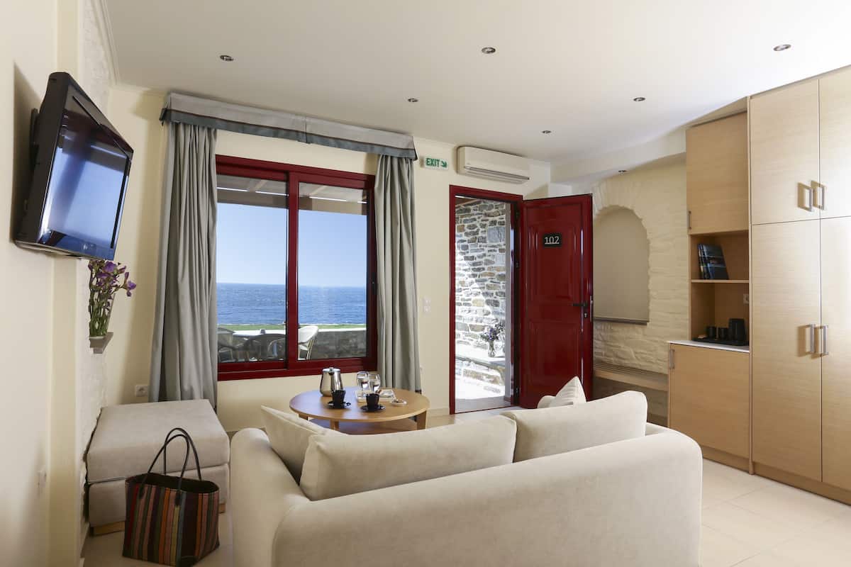 A room with a sofa, a table with a coffee pot and glasses, a TV mounted on the wall, and a view of the sea through a large window.
